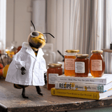 A cartoon bee in a lab coat stands beside jars of honey and cookbooks on a rustic table.