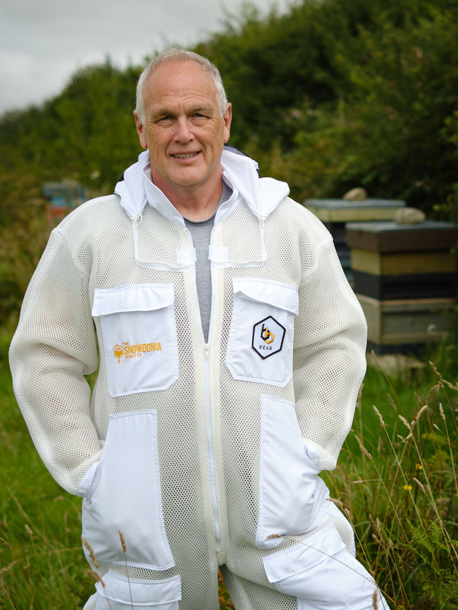 Man wearing a white beekeeping suit in a field with greenery.